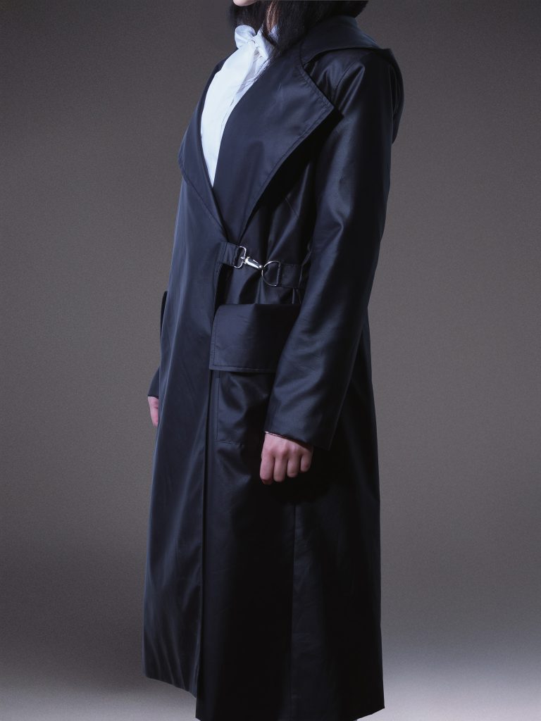 Designer black waterproof trench coat with oversized hood, asymmetric front, side buckle fastening, and contrasting white lining on a light grey studio background.