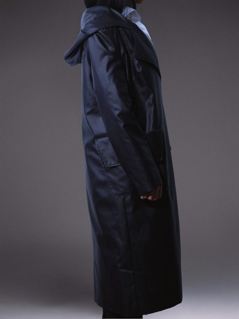 Designer black waterproof trench coat with oversized hood, asymmetric front, side buckle fastening, and contrasting white lining on a light grey studio background.