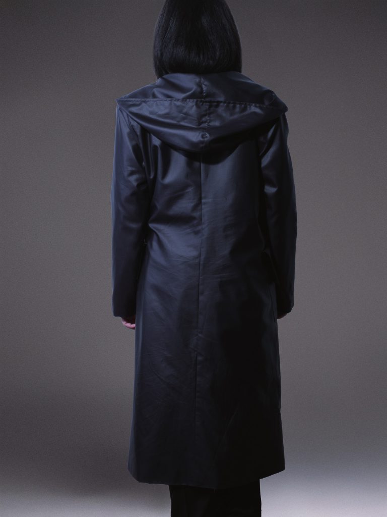 Designer black waterproof trench coat with oversized hood, asymmetric front, side buckle fastening, and contrasting white lining on a light grey studio background.