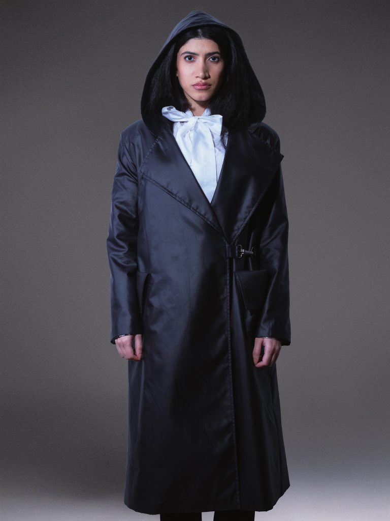 Designer black waterproof trench coat with oversized hood, asymmetric front, side buckle fastening, and contrasting white lining on a light grey studio background.