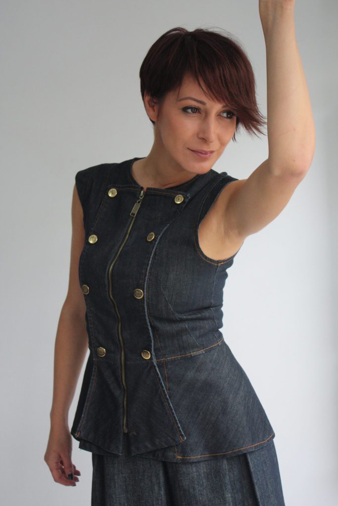 Dark indigo denim peplum vest with front zip, metallic buttons and structured sleeveless silhouette on light grey background