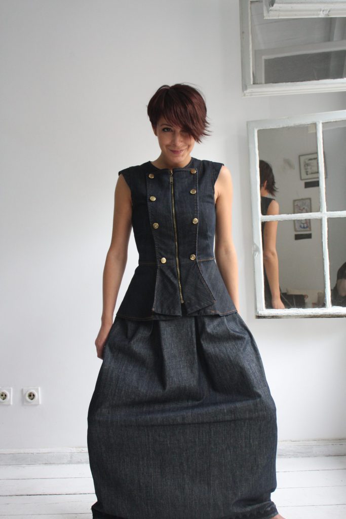 Dark indigo denim peplum vest with front zip, metallic buttons and structured sleeveless silhouette on light grey background